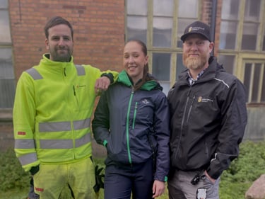 Tre Sekomedlemmar som tittar in i kameran. Två män och en kvinna, de står iklädda arbetskläder från sina företag där de jobbar.